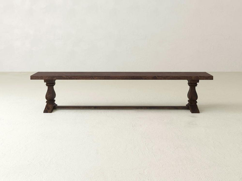 The Heirloom Bench - Tobacco by James+James is crafted from dark wood, featuring a rectangular seat and intricately carved, sturdy legs. A supporting horizontal beam connects the two legs, enhancing its classic and rustic appearance, set against a white background.