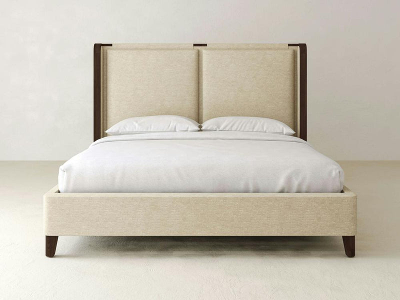 The Holden Bed – Linen by James & James features a luxurious beige upholstered design with a tall, padded headboard and dark solid maple wood accents. It combines elegance and durability with performance fabric, complemented by white bedding and two pillows against a light-colored backdrop.