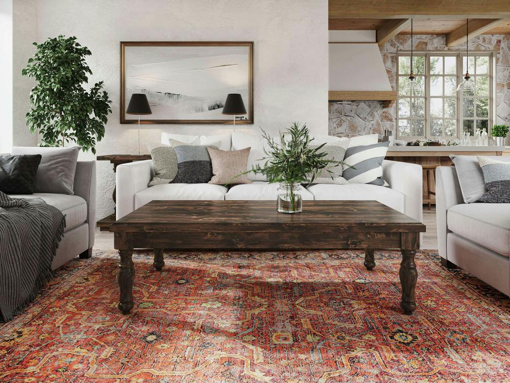 A cozy living room featuring a white sofa adorned with gray and white pillows, complemented by the Abigail Coffee Table in Tobacco by James+James, decorated with a plant in a glass vase. A vibrant red patterned rug adds color to the scene. The back wall showcases a large framed artwork flanked by two wall lamps and a potted plant. In the background, you can see the rustic kitchen design.