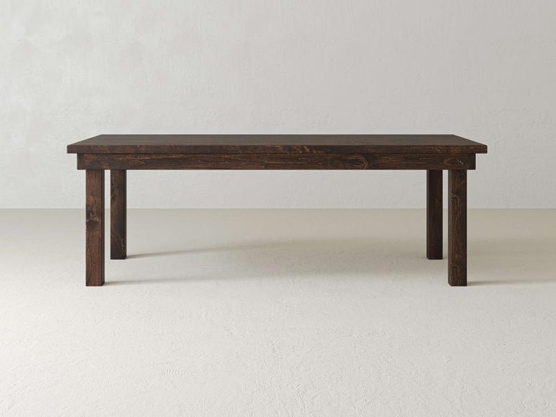 A James+James Farmhouse Dining Table in Tobacco finish, featuring a simple dark wooden design with a rectangular top and four sturdy legs, stands on a light-colored floor against a plain white wall.